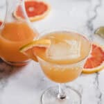A cocktail glass with a grapefruit drink, a large ice cube, and a grapefruit wedge garnish is on a marble surface. A carafe with more drink and grapefruit slices are in the background.