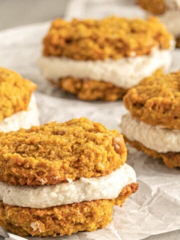 Pumpkin whoopie pies with cream filling are arranged on crinkled parchment paper. The cookies are round, orange-brown, and sandwiched with white cream, set on a light wooden surface.
