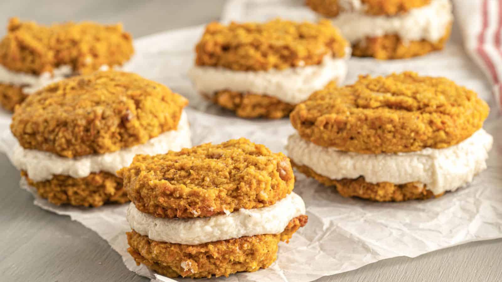 Pumpkin whoopie pies with cream filling are arranged on crinkled parchment paper. The cookies are round, orange-brown, and sandwiched with white cream, set on a light wooden surface.