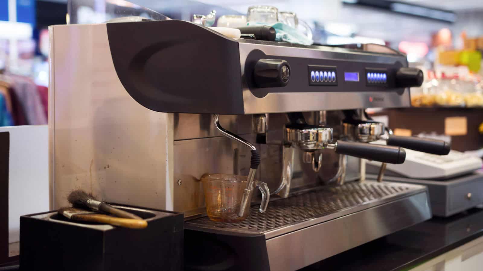 A stainless steel commercial espresso machine with dual group heads and steam wands is on a counter. Nearby are cleaning brushes and a small glass. The environment appears to be a caf&eacute; or coffee shop setting.
