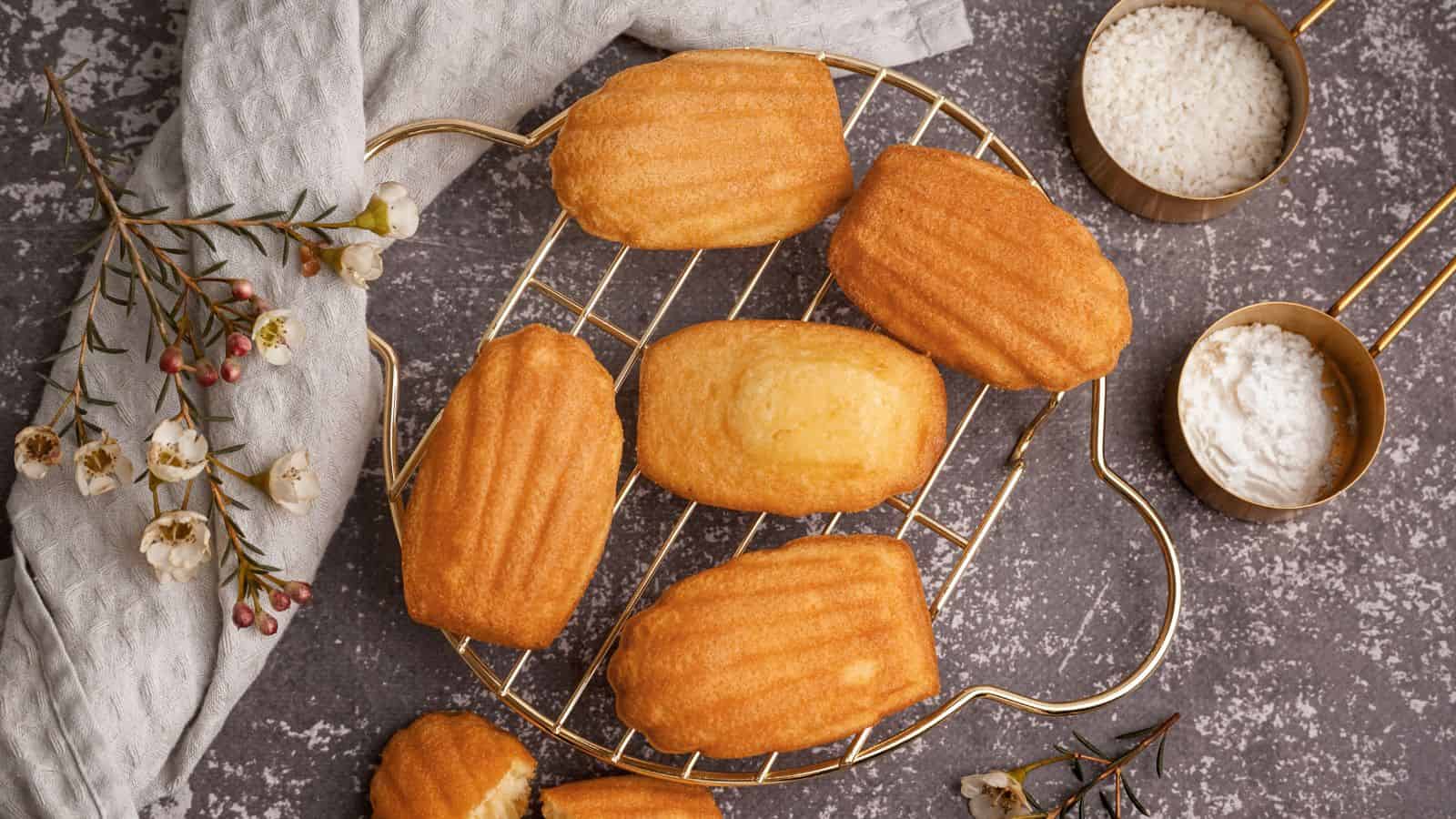 Golden-brown madeleines are arranged on a wire cooling rack, set on a dark countertop. To the right, two small bowls contain powdered sugar and grated coconut. A light gray cloth and some small flowers are on the left.