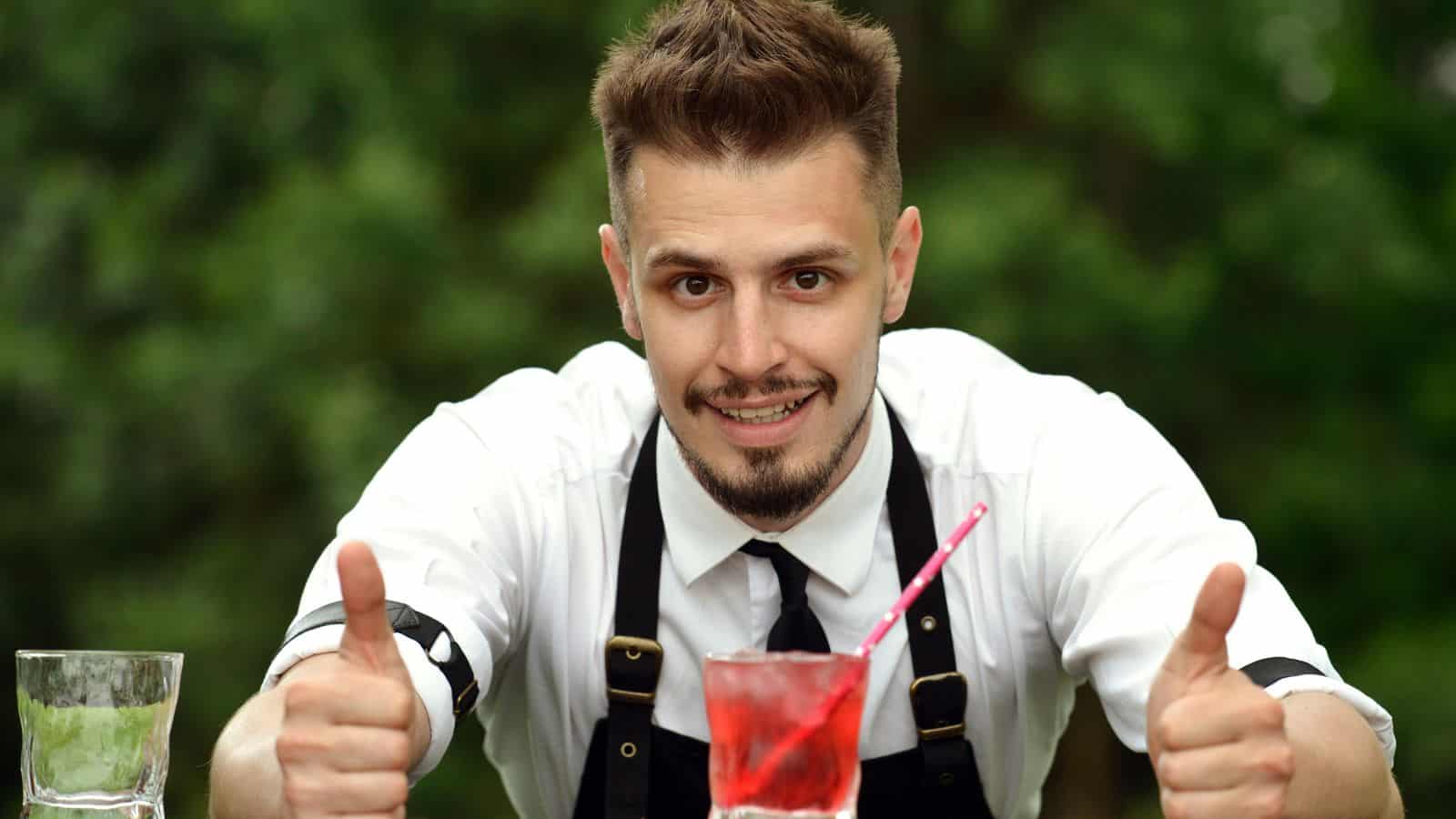 A person wearing a white shirt, black apron, and tie gives two thumbs up with a smile. In front are two glass drinks: one empty and one with a red liquid and a straw. The background shows green foliage.