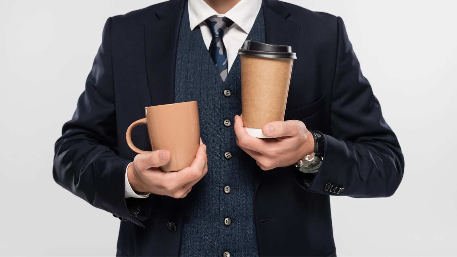 A person in a dark suit holds a brown ceramic mug in their left hand and a brown paper coffee cup with a black lid in their right hand. The background is plain white.