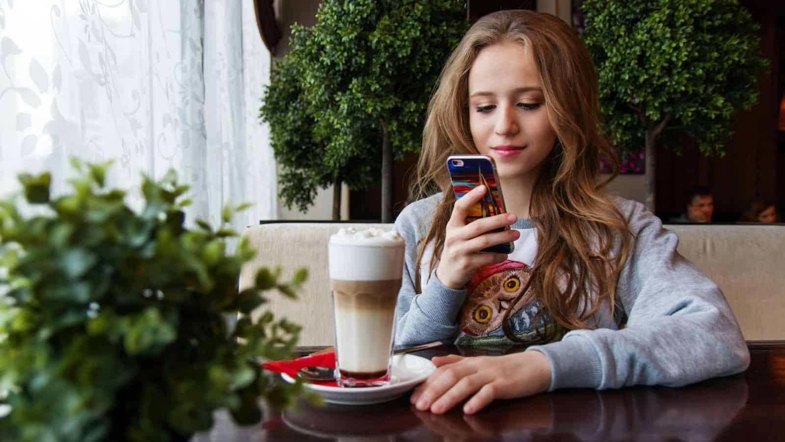 A woman with long hair sits at a table in a cafe, looking at her phone. In front of her is a latte with foam in a glass on a saucer. There are two small green potted plants nearby. She wears a sweatshirt with a colorful owl design.