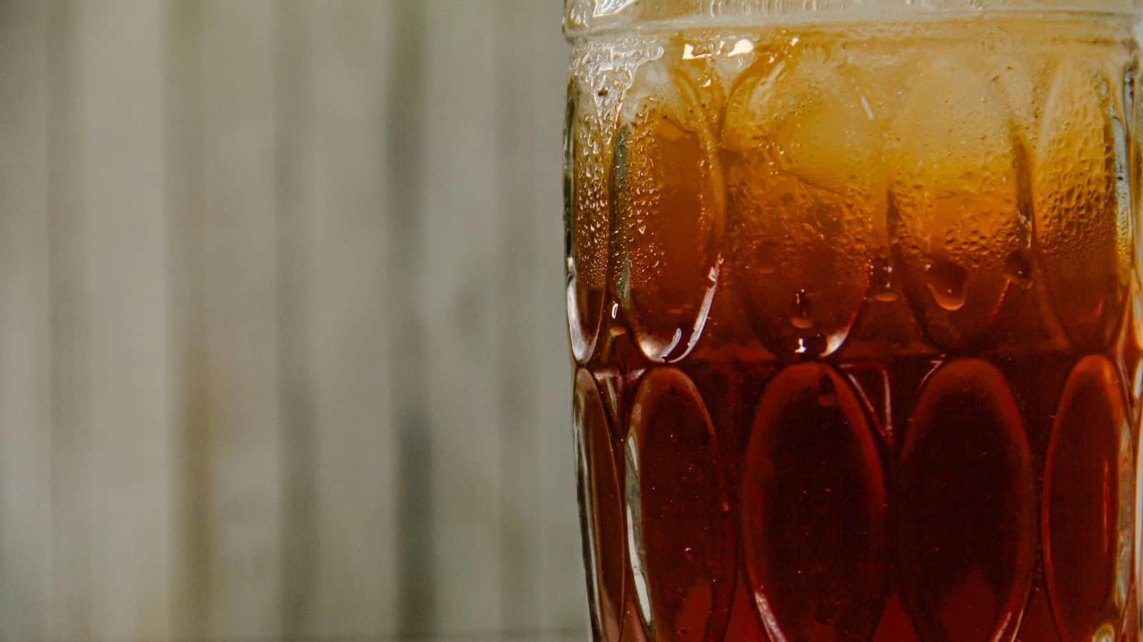 Close-up of a glass filled with iced tea. The glass has a textured pattern and contains ice cubes. The liquid is a dark amber color. The background is blurred with vertical wooden panels.