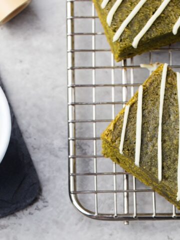 A white cup filled with frothy green matcha sits on a dark slate coaster. Next to it, three square pieces of matcha bread with white icing drizzle are placed on a metal cooling rack, all on a light gray surface.