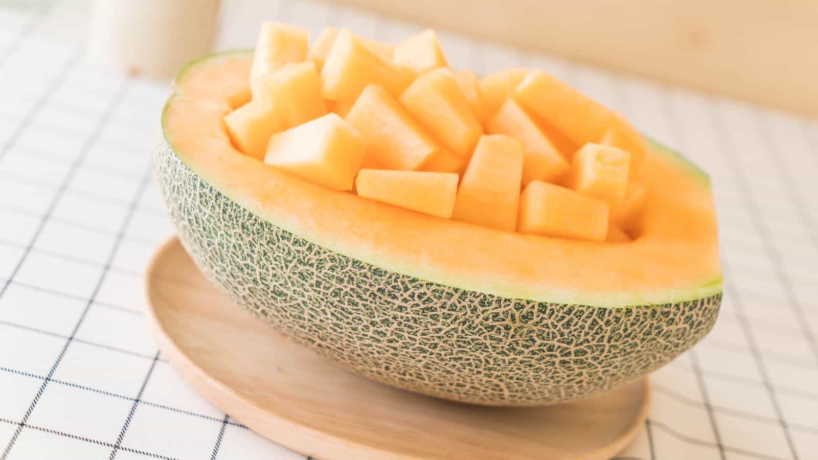 A halved cantaloupe with seeds removed, filled with cubed pieces of its own orange flesh. The fruit is on a wooden plate resting on a checkered tablecloth.