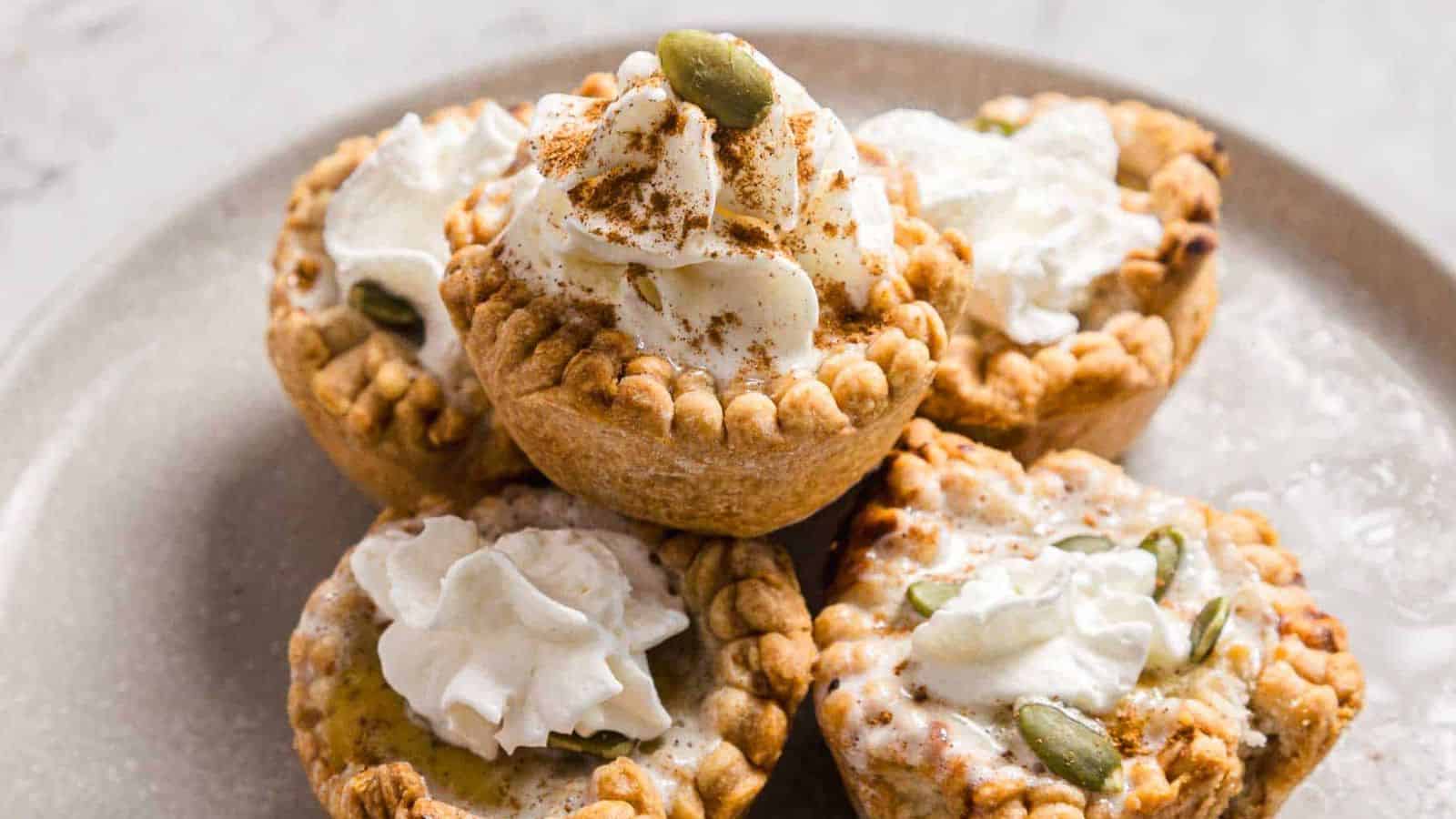 A plate of five mini pies topped with whipped cream and sprinkled with cinnamon. Some have pumpkin seeds as garnish. The pies have a golden-brown crust and are arranged in a pyramid shape on the plate.