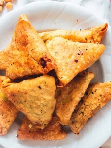 A plate of golden-brown triangular samosas is placed on a marble surface. Surrounding the plate are scattered chickpeas, fresh cilantro, a red pepper, and a cutting board corner. The samosas appear crispy and seasoned.