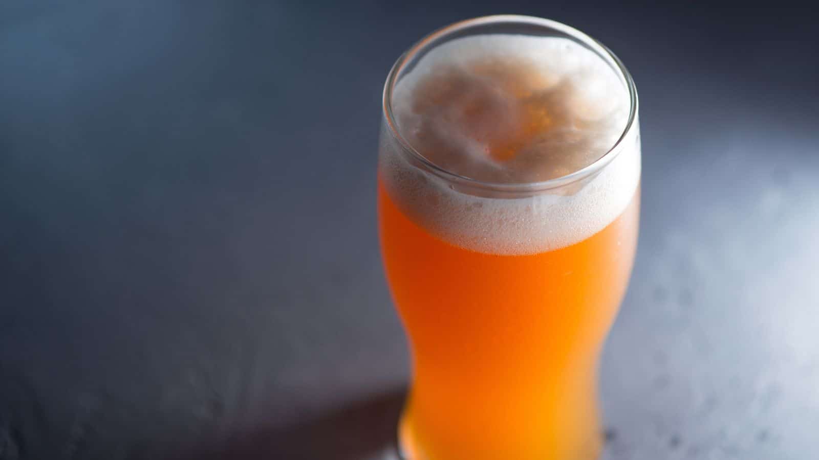 A glass of orange-hued beer with a frothy head sits on a dark, textured surface. The lighting highlights the beer's color and bubbles, creating a slight contrast against the background.