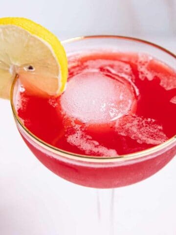 A cocktail glass filled with a bright red drink and a large ice sphere, garnished with a lemon slice on the rim. A halved lemon and a cherry are visible in the background on a light surface.