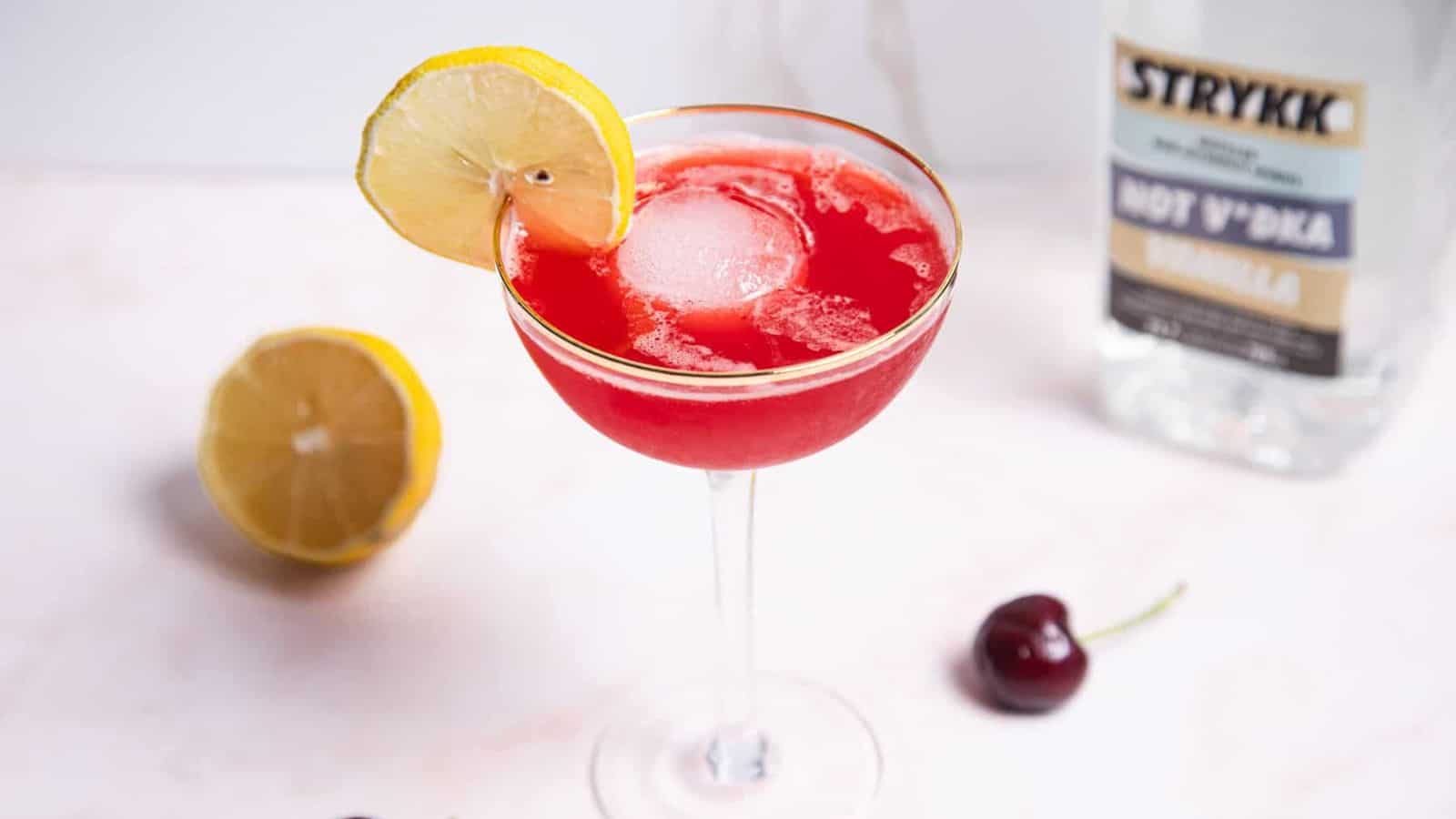 A mocktail in a glass with a gold rim is filled with a red liquid and a large round ice cube. A lemon slice is on the rim. A halved lemon and a cherry are nearby on a white surface.
