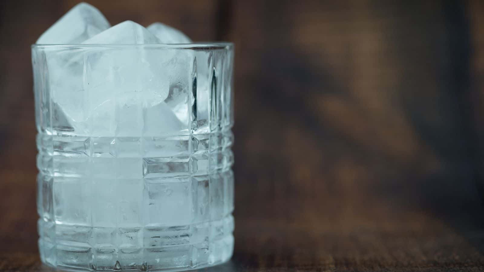 A clear glass tumbler filled with large ice cubes sits on a wooden surface. The background is blurred, creating contrast between the sharp lines of the glass and ice and the muted colors behind.