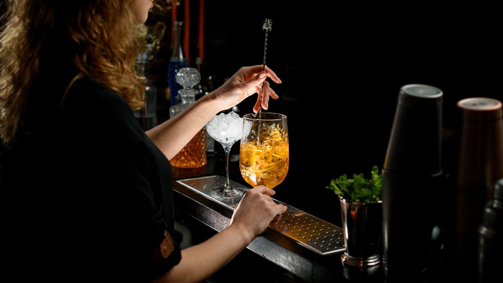 A bartender is stirring an orange cocktail in a tall glass with a straw, surrounded by a shaker, ice, and a glass container with green herbs. The setting is dimly lit, emphasizing the drink's vibrant color.