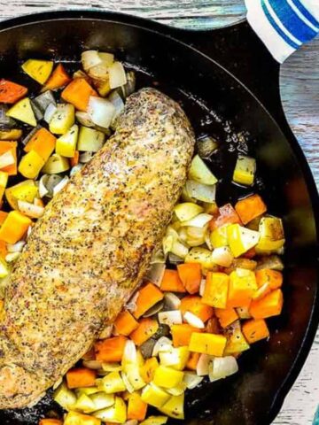 A seasoned fish fillet is placed in a cast iron skillet, surrounded by roasted cubes of potatoes, carrots, and other vegetables. The skillet rests on a wooden surface next to a blue-striped cloth.