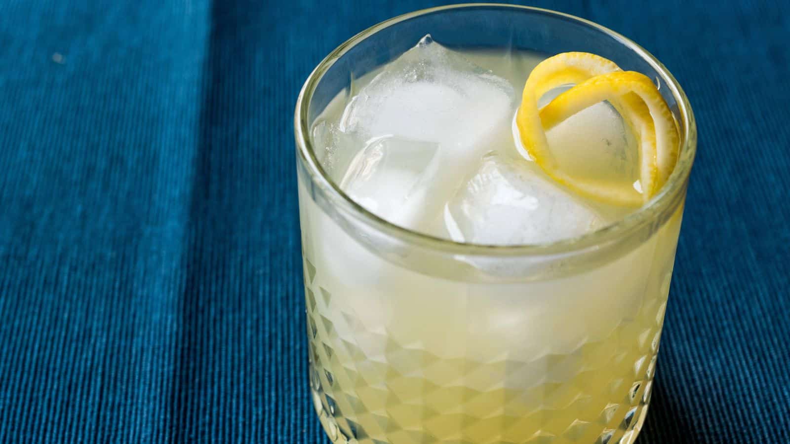 A glass of light-colored cocktail sits on a blue cloth surface. The drink contains ice cubes and is garnished with a twist of lemon peel. The glass features a textured pattern.