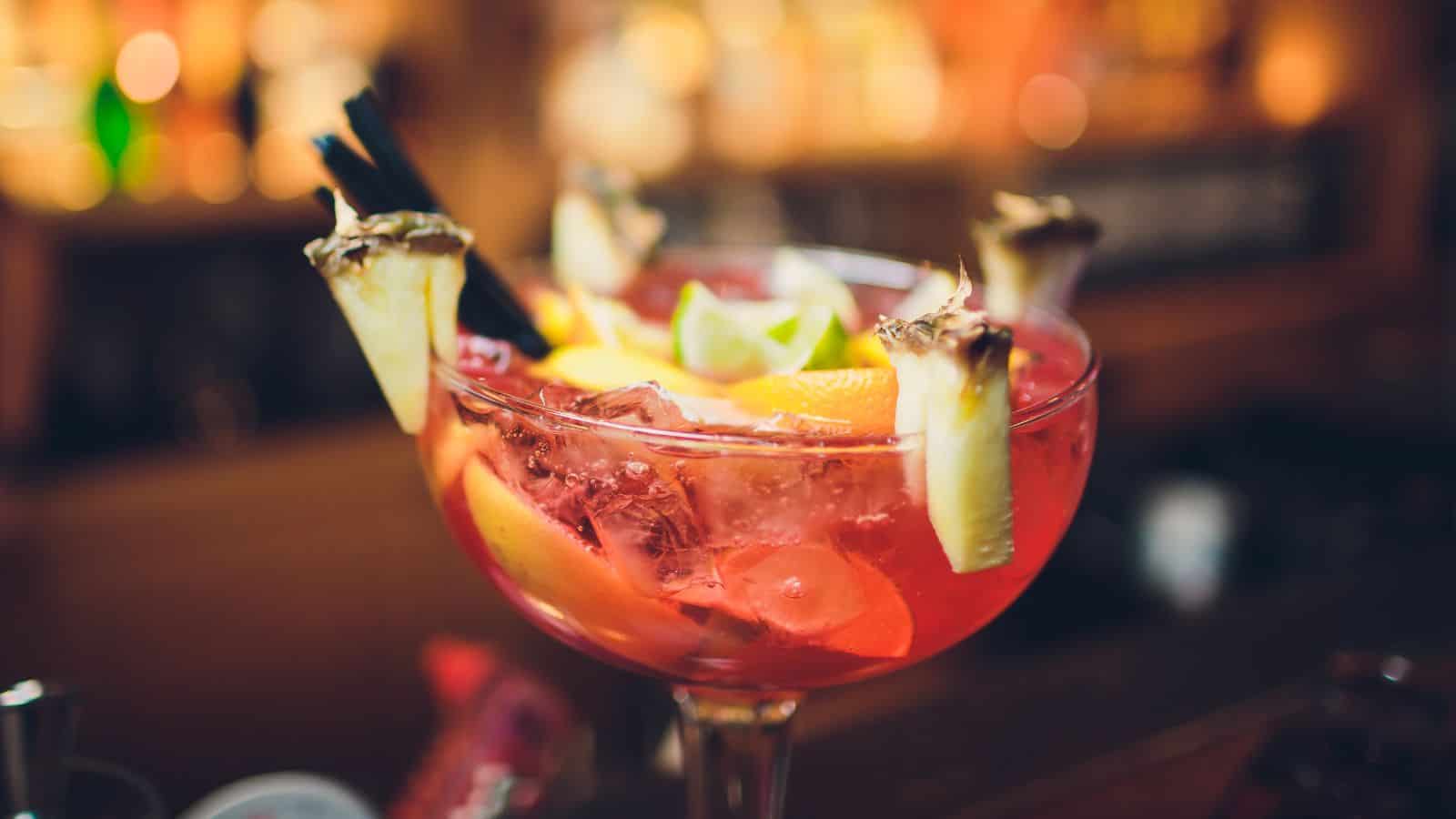 A large cocktail glass filled with a red drink, ice, and garnished with pineapple slices, orange wedges, and lime twists. Two black straws are inserted. The background is blurred, suggesting a bar setting.