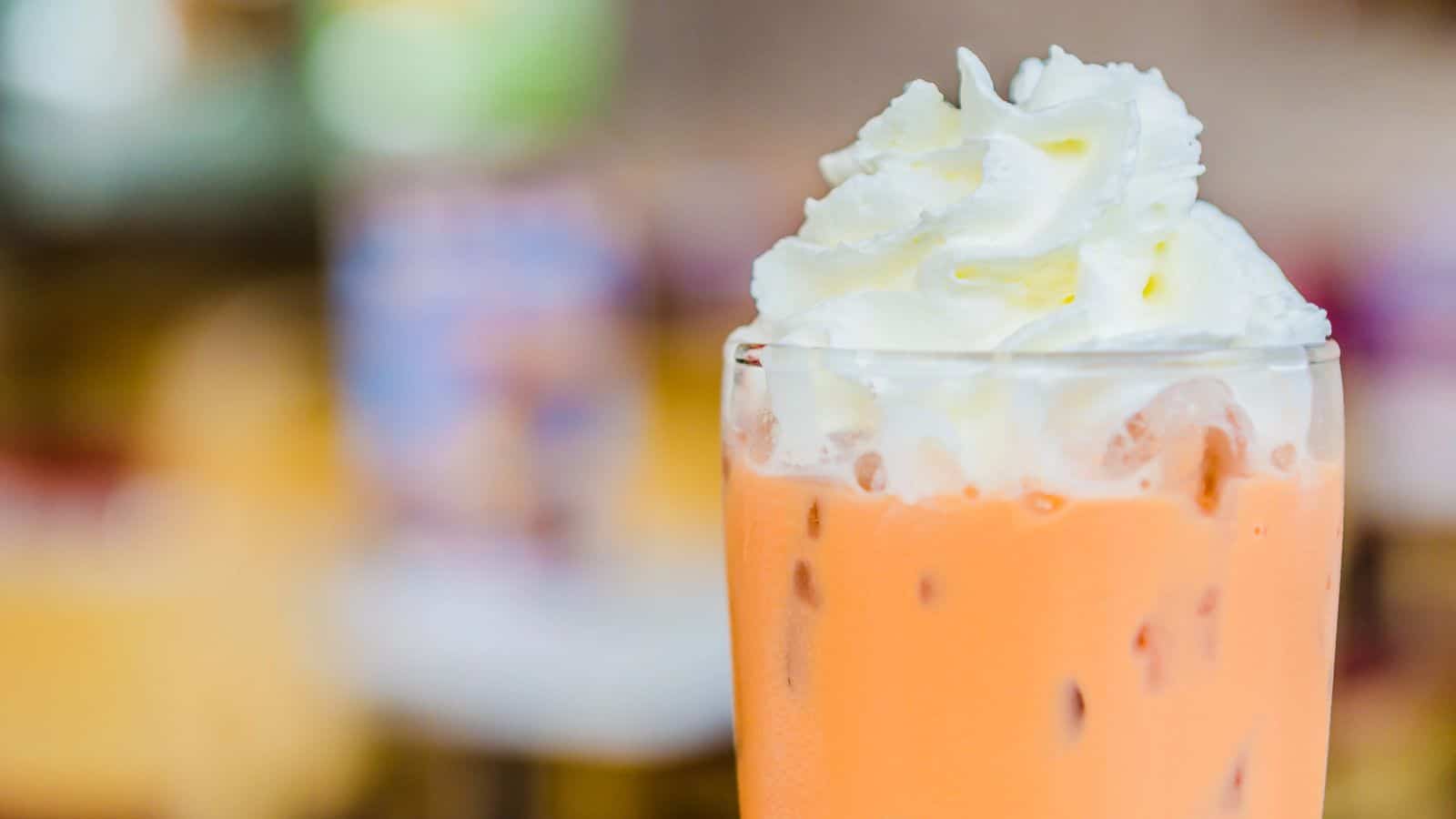 A close-up of a glass filled with iced Thai tea topped with a generous swirl of whipped cream. The beverage has a bright orange color, and the background is blurred, emphasizing the focus on the drink.