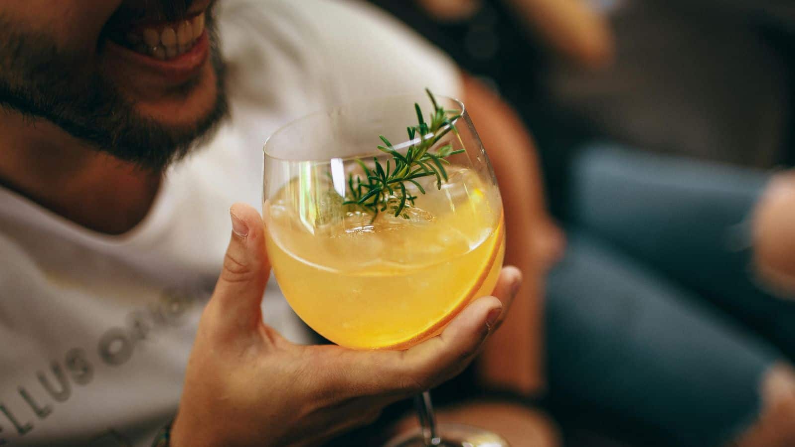 A person holds a cocktail in a large glass, garnished with a sprig of rosemary and an orange slice. The drink has a light orange hue. The person's face is partially visible, showing a smile. The background is blurred.