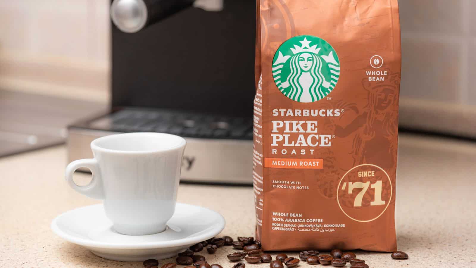 Bag of Starbucks Pike Place medium roast coffee displayed on a kitchen counter next to a white espresso cup and saucer. Coffee beans are scattered nearby, and a portion of a coffee machine is visible in the background.