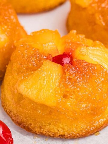 Mini pineapple upside-down cakes are topped with pineapple pieces and maraschino cherries. They are golden brown, arranged on a white surface, with extra cherries nearby.