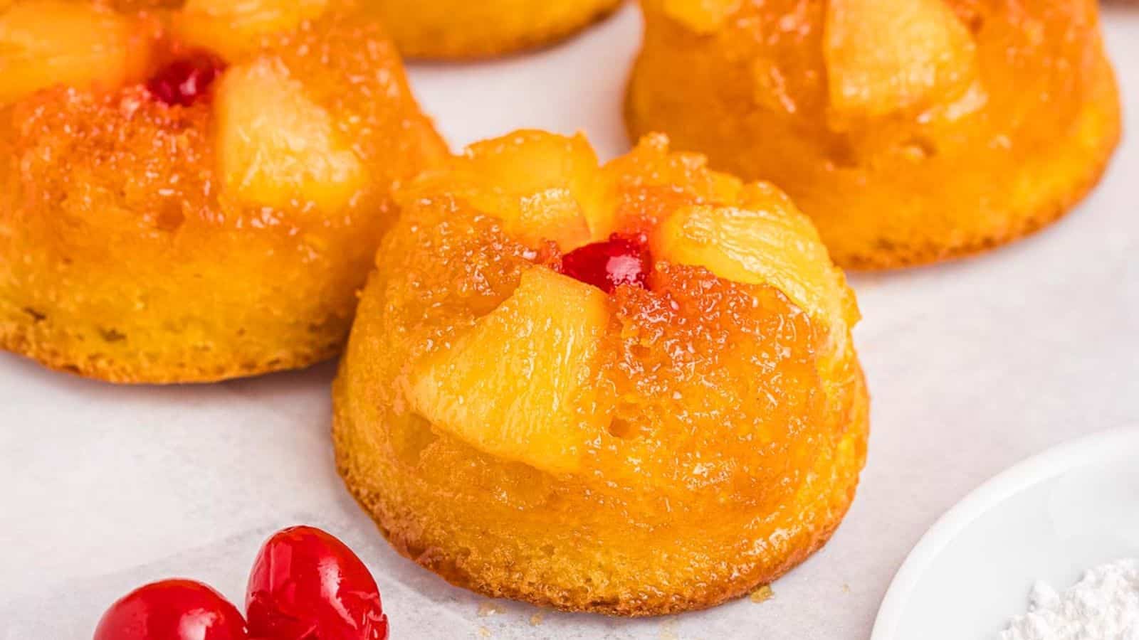 Mini pineapple upside-down cakes topped with pineapple chunks and cherries on a white surface.