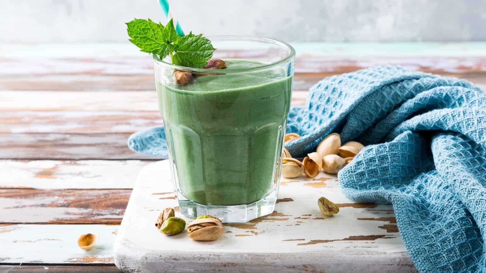 A glass of green smoothie garnished with a mint leaf and pistachios sits on a white cutting board. A blue cloth and scattered pistachios surround the glass. The background features a wooden surface with a light blue and white finish.