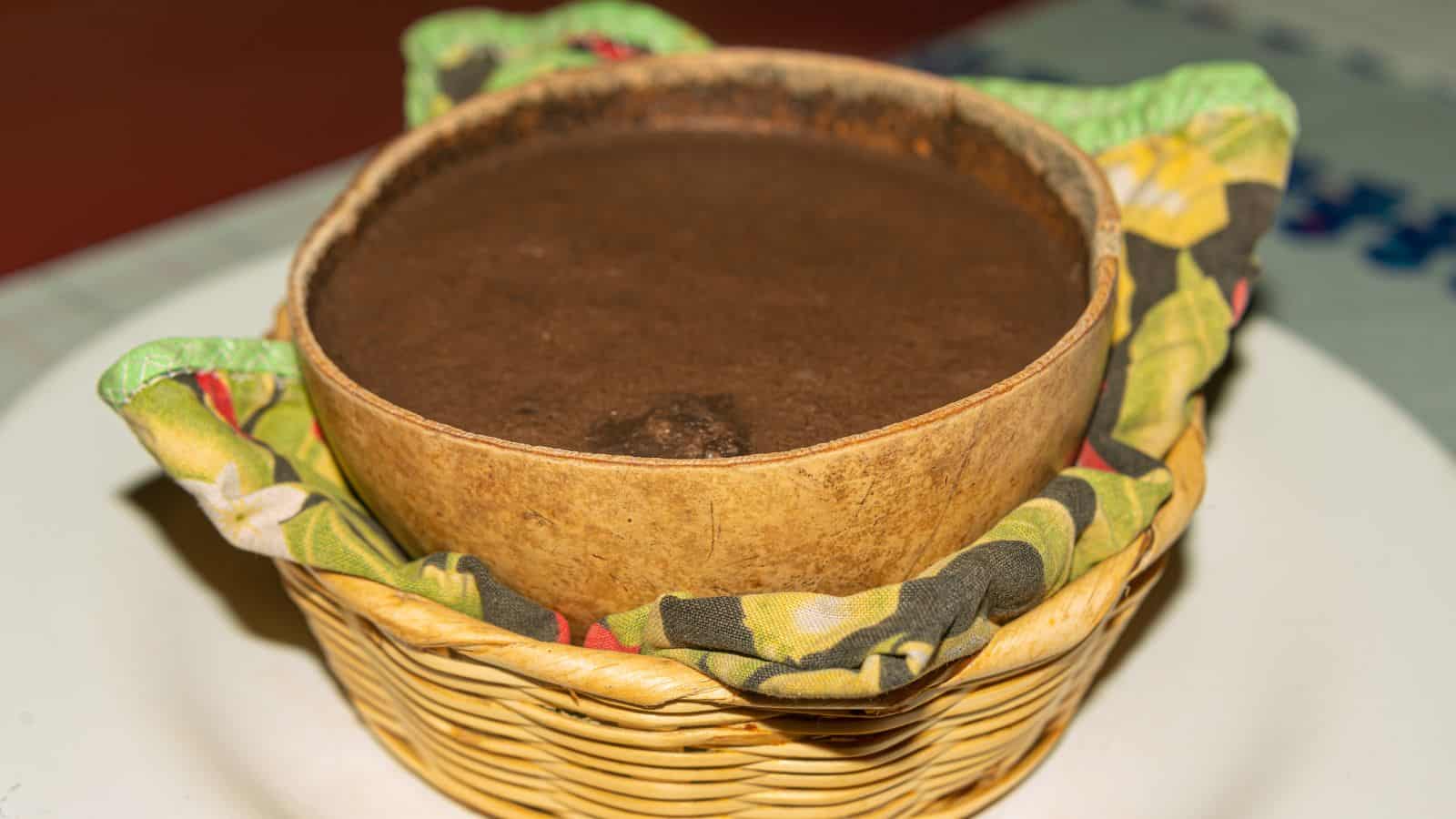 A brown liquid in a bowl rests on a colorful cloth inside a wicker basket. The setting is on a table with a checkered cloth. The overall appearance suggests a traditional or homemade beverage or dish.