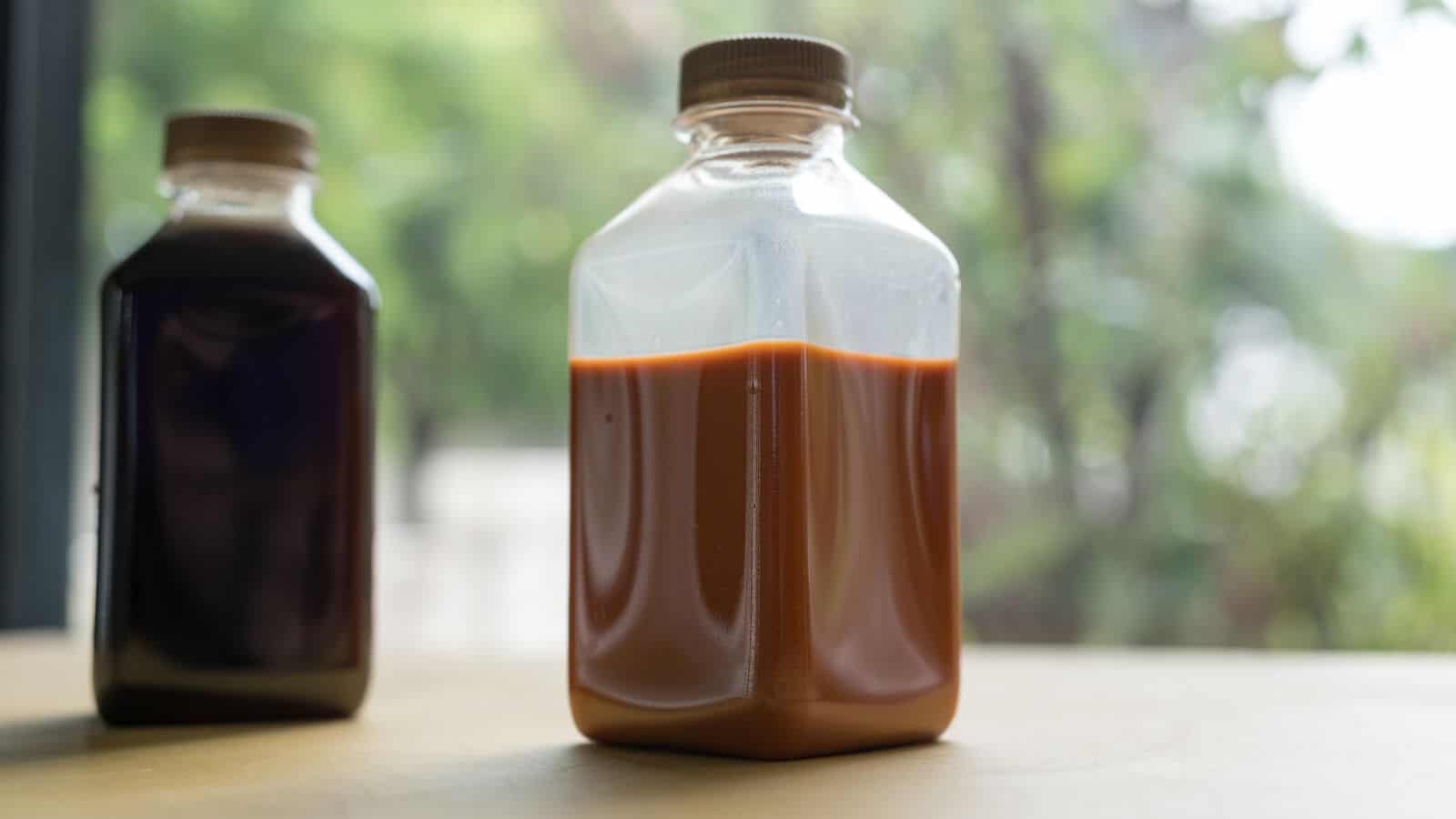 Two plastic bottles with screw-on caps sit on a surface. The closer bottle contains an orange-brown liquid, while the bottle in the background holds a darker liquid. Blurred greenery is visible through a window in the background.