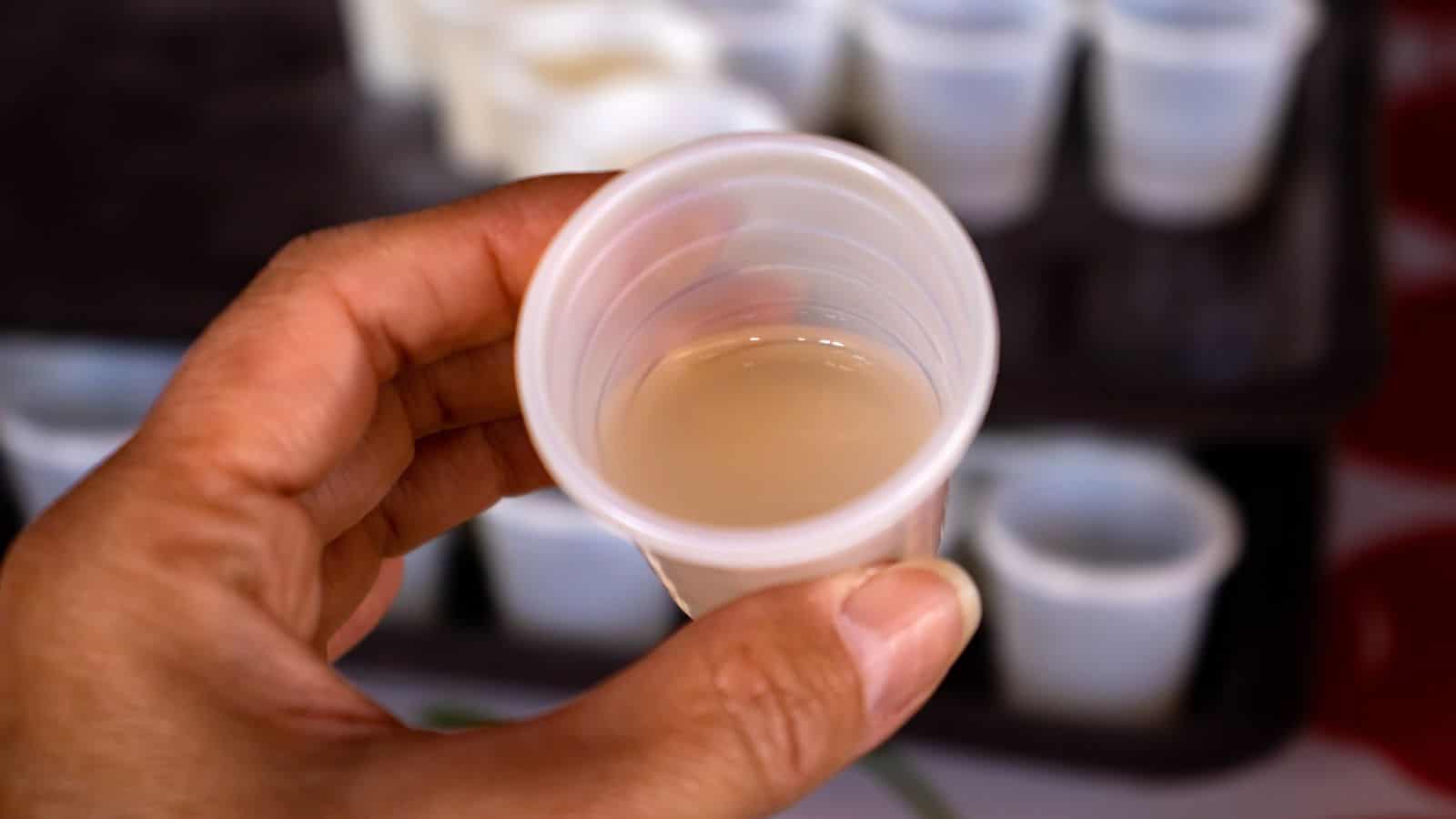 A hand holds a small, translucent plastic cup containing a beige liquid. Several similar cups are blurred in the background on a tray.