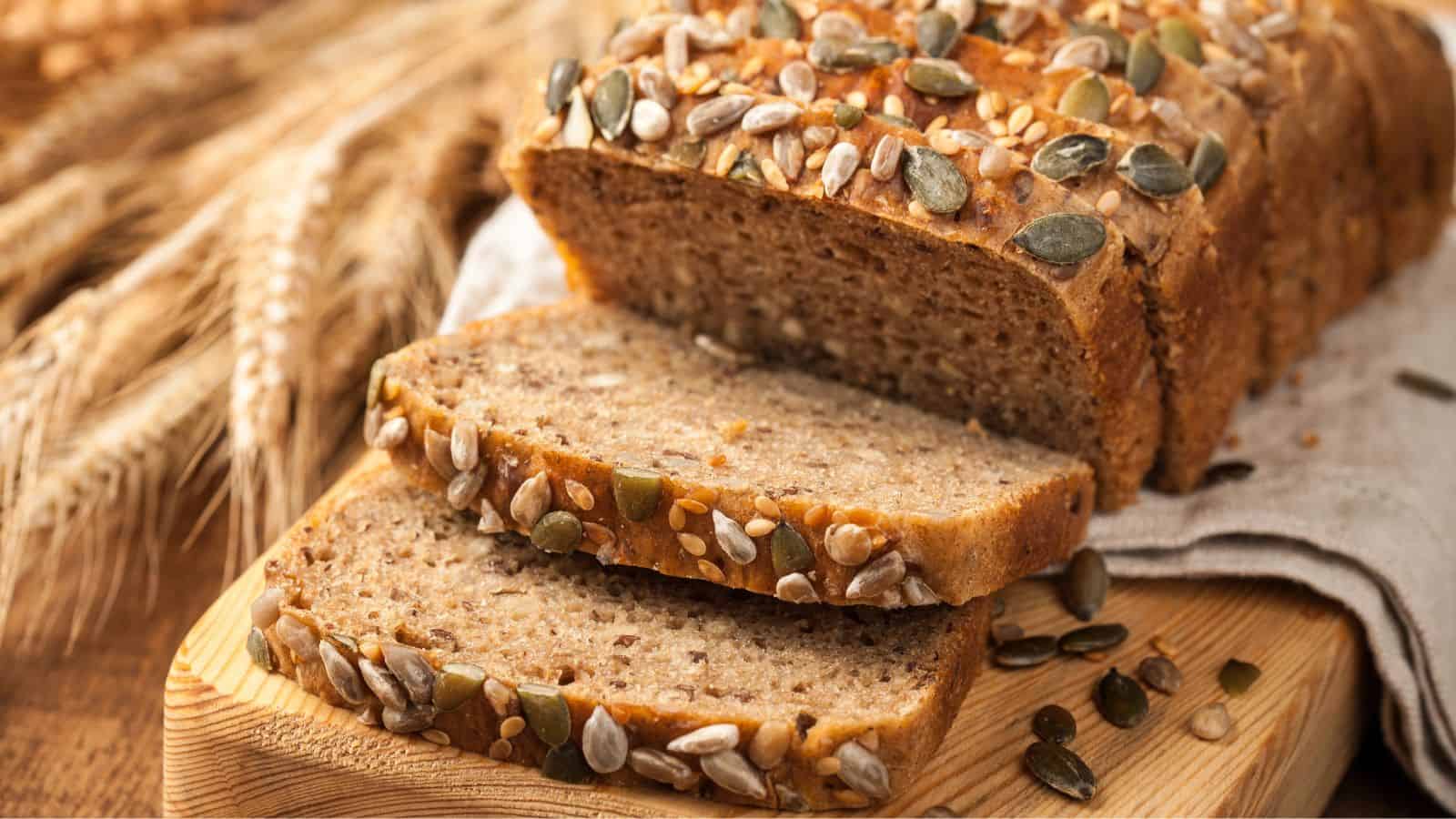 Sliced loaf of multigrain bread topped with a variety of seeds, placed on a wooden cutting board. Wheat stalks are visible in the background, partially out of focus.