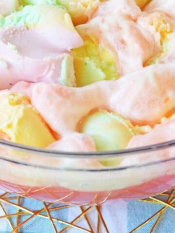 A glass bowl filled with a fizzy, colorful punch containing scoops of pastel-colored sherbet. The bowl sits on a blue and white checkered cloth with a gold wire tray underneath. Part of a similar drink in a glass is visible on the side.