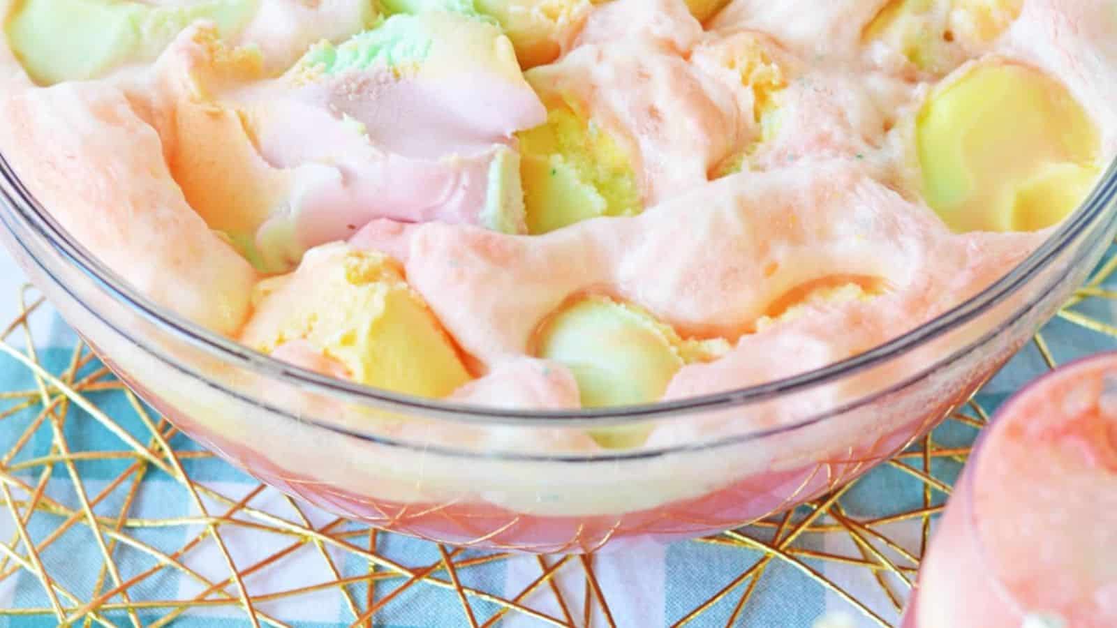 A glass bowl filled with a fizzy, colorful punch containing scoops of pastel-colored sherbet. The bowl sits on a blue and white checkered cloth with a gold wire tray underneath. Part of a similar drink in a glass is visible on the side.