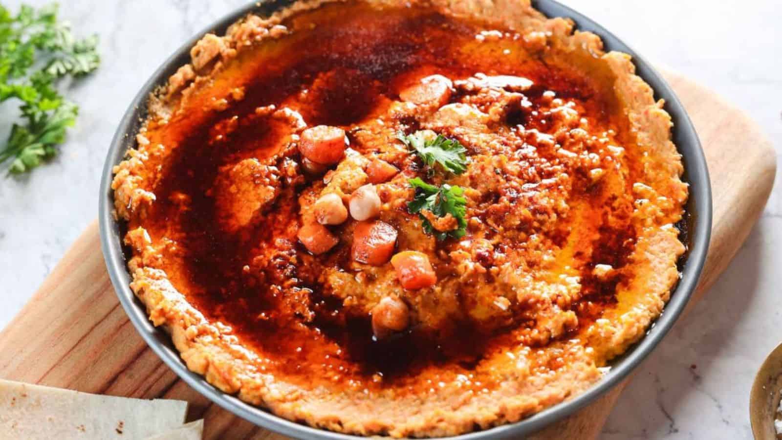 A bowl of hummus topped with spicy red sauce and pieces of chickpeas and carrots, garnished with parsley. The bowl is placed on a wooden board with a partially visible sprig of herbs and flatbread nearby.