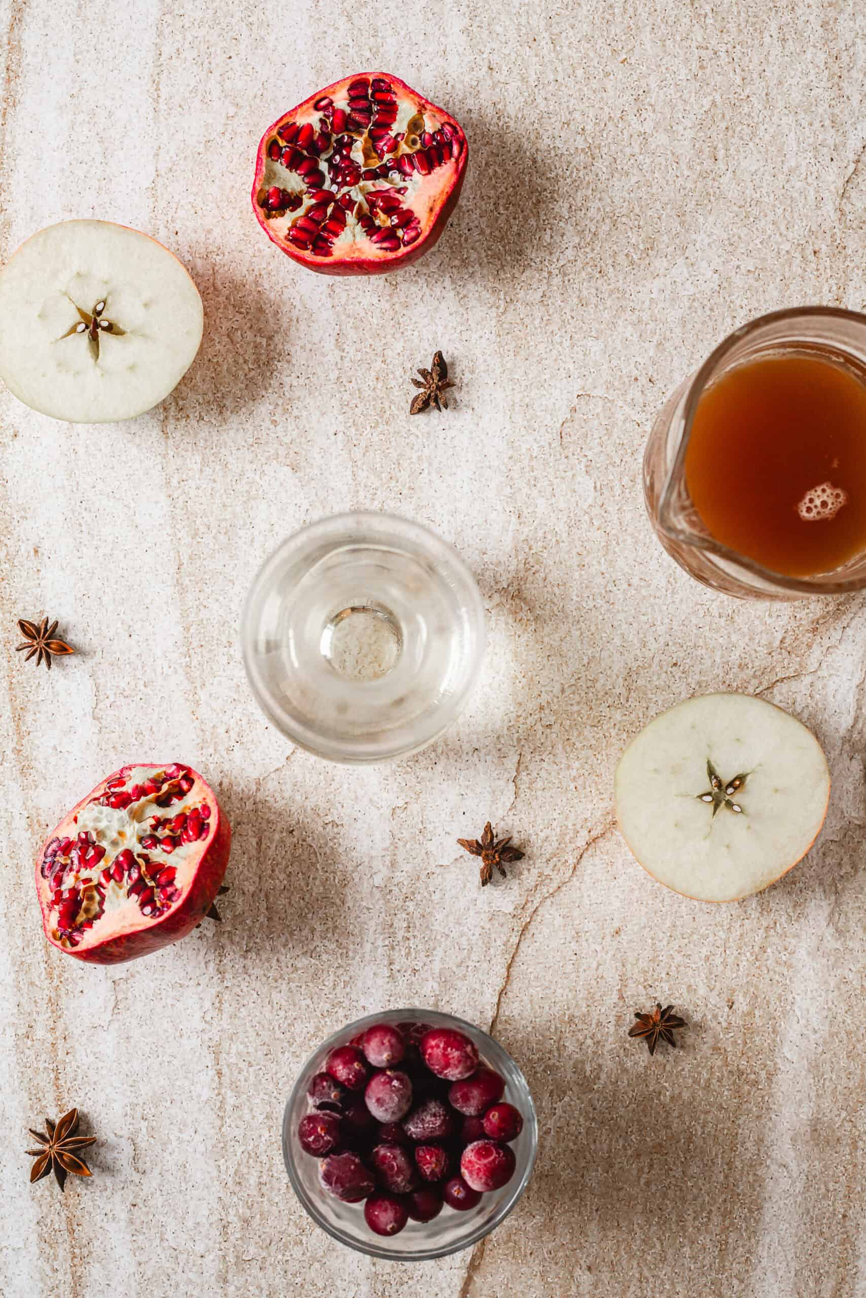 A flat lay of a halved pomegranate, halved apple, a glass of juice, a glass of clear liquid, a bowl of cranberries, and scattered star anise on a light textured surface.