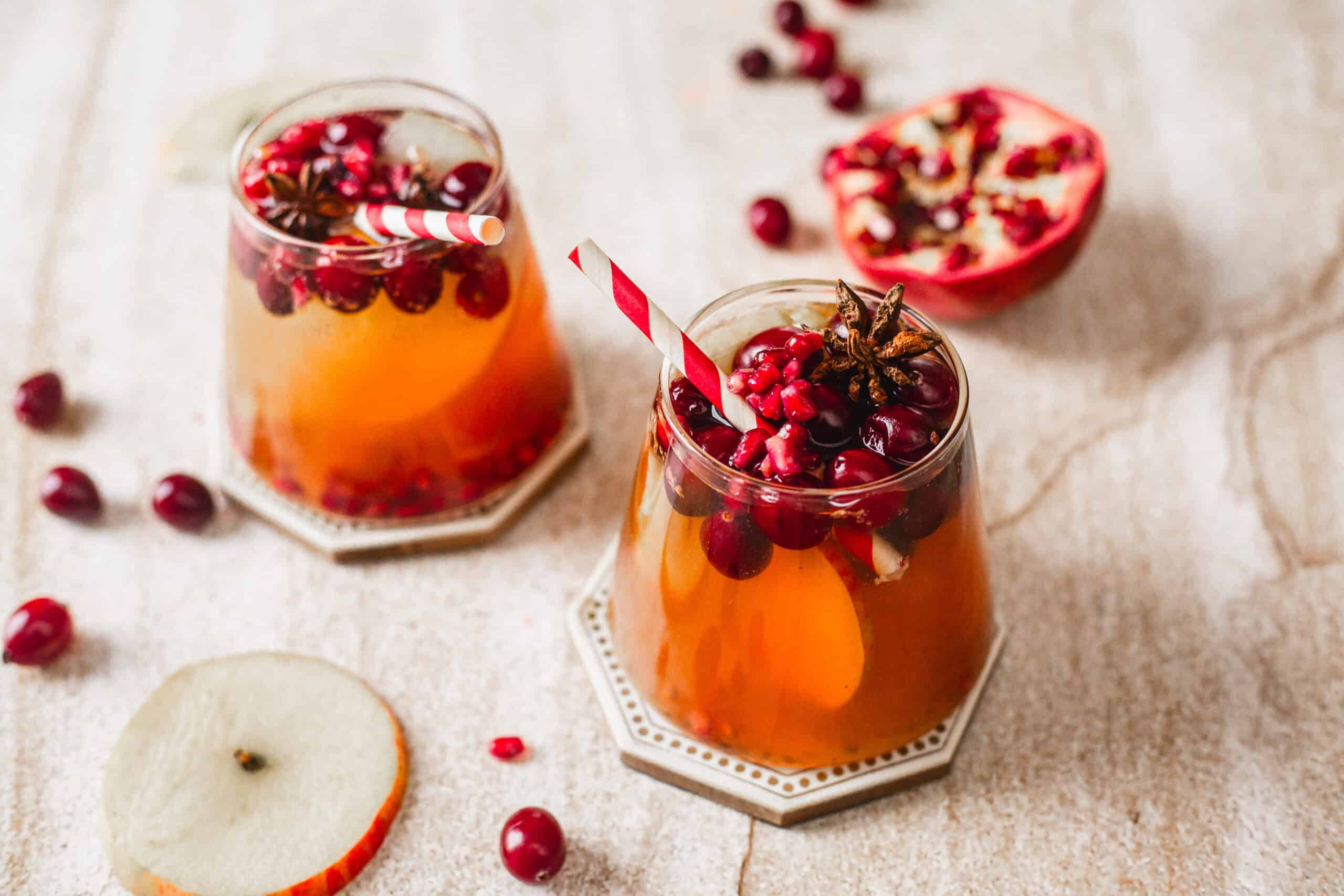Two glasses of a festive drink sit on a textured surface. The glasses are garnished with cranberries, pomegranate seeds, and star anise. Each glass has a striped straw. Slices of apple and loose cranberries are scattered around.