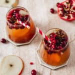 Two glass cups filled with an orange-hued drink, topped with cranberries, pomegranate seeds, and star anise, sit on hexagonal coasters. Red and white striped straws are in the glasses. Slices of apple, pomegranate, and cranberries surround them.
