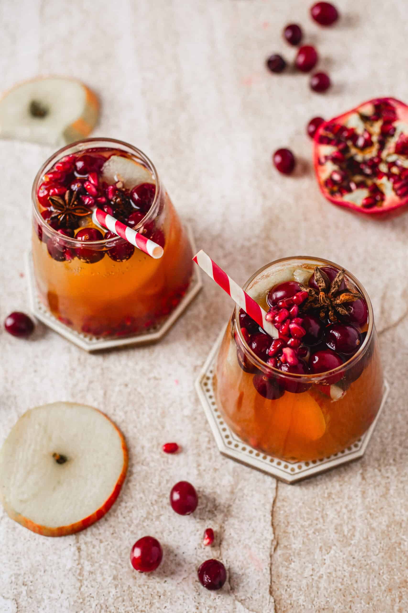 Two glass cups filled with an orange-hued drink, topped with cranberries, pomegranate seeds, and star anise, sit on hexagonal coasters. Red and white striped straws are in the glasses. Slices of apple, pomegranate, and cranberries surround them.