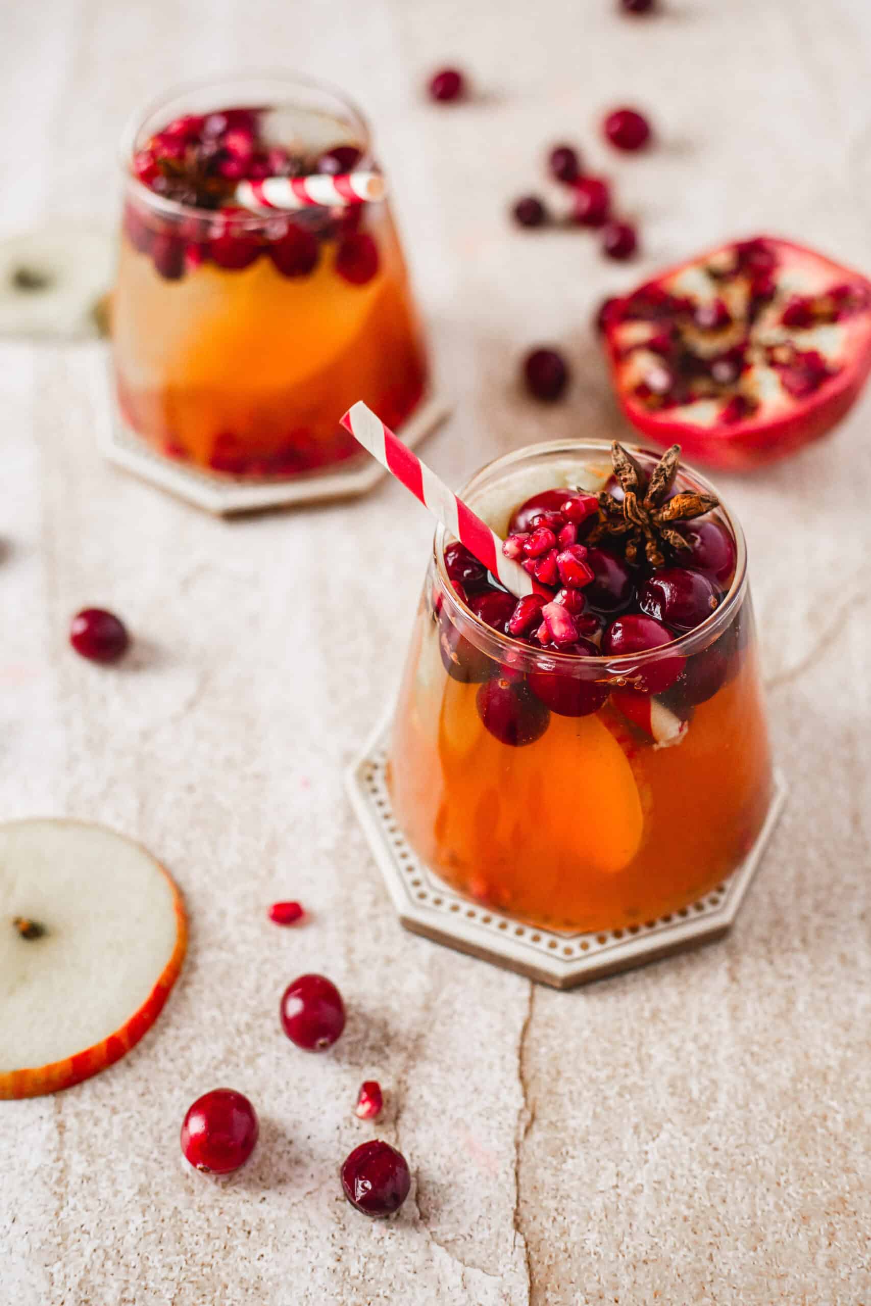Two glasses of a festive beverage garnished with cranberries, pomegranate seeds, and star anise are on a textured surface. A red-and-white straw is in one glass. Nearby are scattered cranberries, pomegranate seeds, and apple slices.