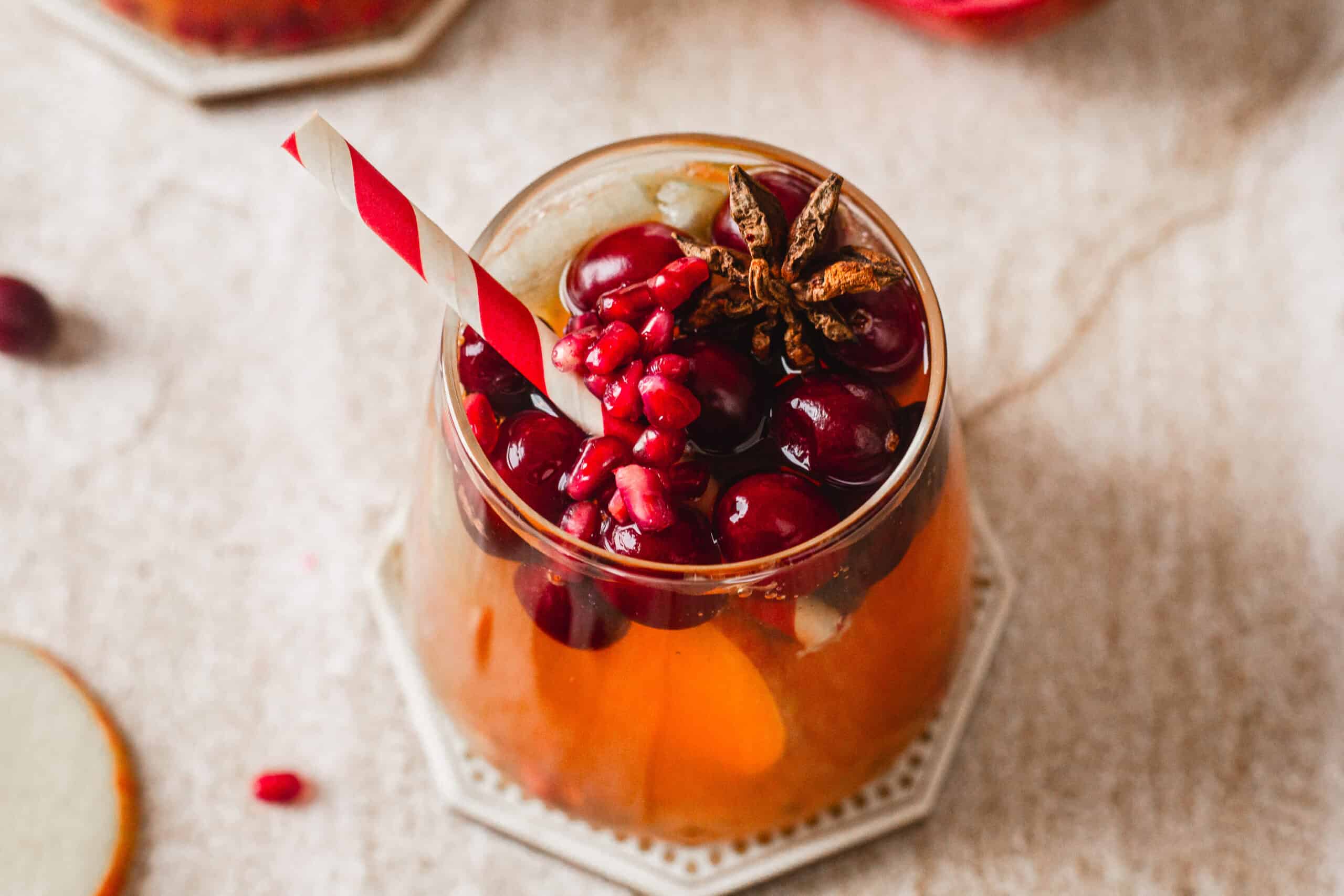 A glass filled with a reddish-orange drink, garnished with pomegranate seeds, cranberries, and a star anise. A red and white striped straw is also in the glass, resting on a hexagonal coaster.