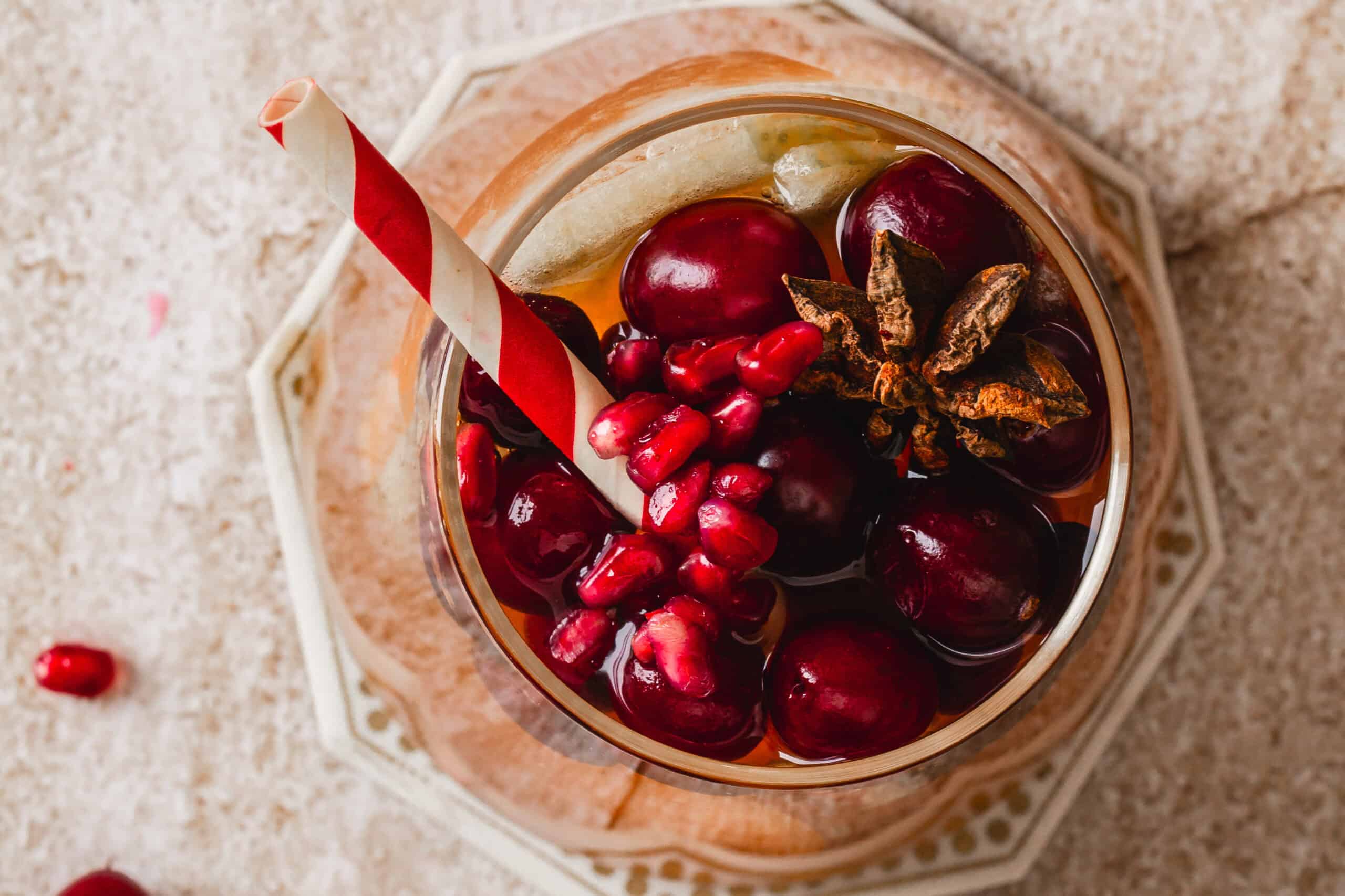 A top view of a glass filled with a drink containing whole cranberries, pomegranate seeds, and star anise. The glass has a red and white striped straw. It is placed on a detailed coaster on a textured surface.