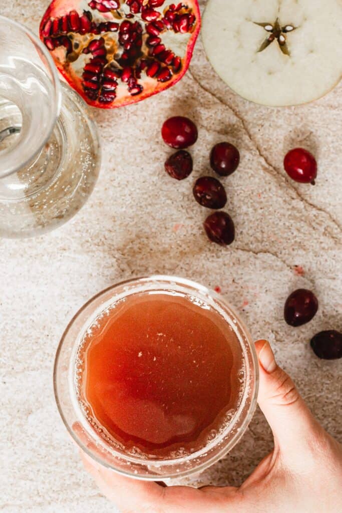 A hand holds a glass filled with a reddish-brown liquid. Nearby, a glass pitcher sits beside pomegranate seeds, halved pomegranate, halved apple, and scattered cranberries on a light stone surface.