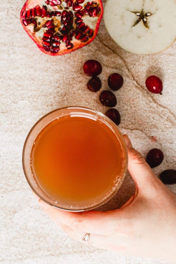 A hand holds a glass of brownish-red juice. In the background, there is a halved pomegranate, an apple slice, and several cranberries scattered on a textured surface.