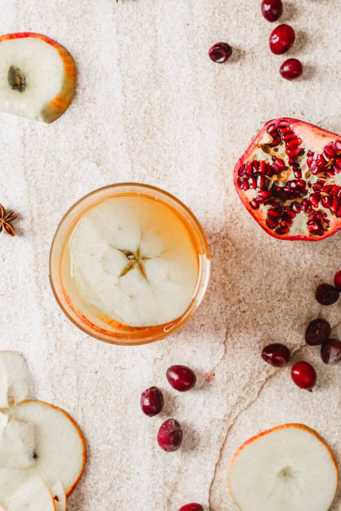 A glass of apple juice is garnished with a slice of apple on a textured surface. Surrounding the glass are apple slices, cranberries, a half-cut pomegranate, and star anise.