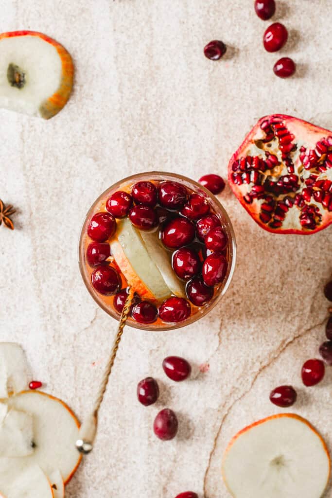 Top view of a beverage garnished with sliced apples and cranberries in a glass with a metal stirrer. Nearby are a halved pomegranate, apple slices, and scattered cranberries on a textured surface.