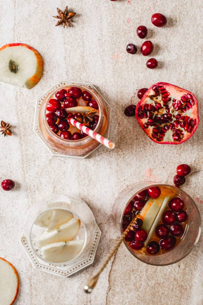 Top view of two drinks in glasses, each with apple slices and cranberries. One glass has a striped straw. Nearby are scattered cranberries, a halved pomegranate, apple slices, and star anise on a textured surface.