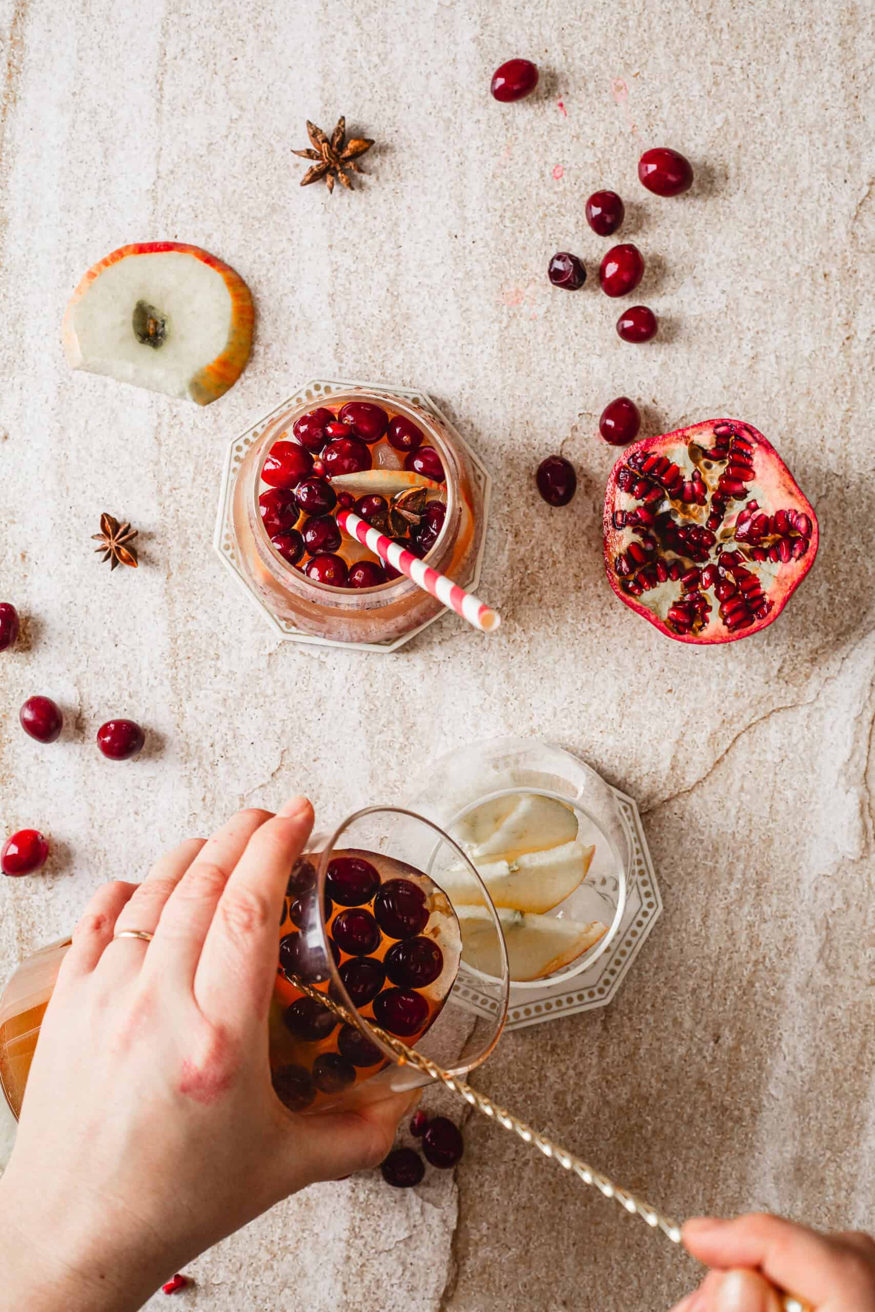 Hands preparing a beverage in a glass with cranberries and a straw, surrounded by another glass with apple slices, a pomegranate half, cranberries, apple slice, star anise, and a light textured surface.