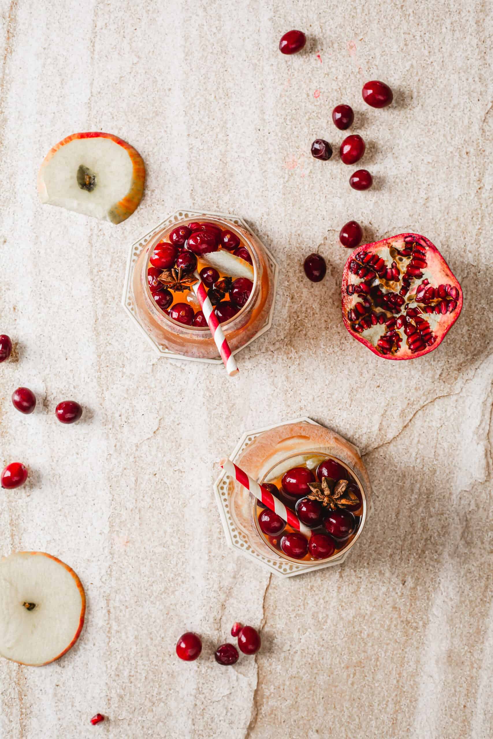 Two glasses with red straws contain a drink garnished with cranberries and pomegranate seeds, placed on a light stone surface. Surrounding them are scattered cranberries, a half pomegranate, and apple slices.
