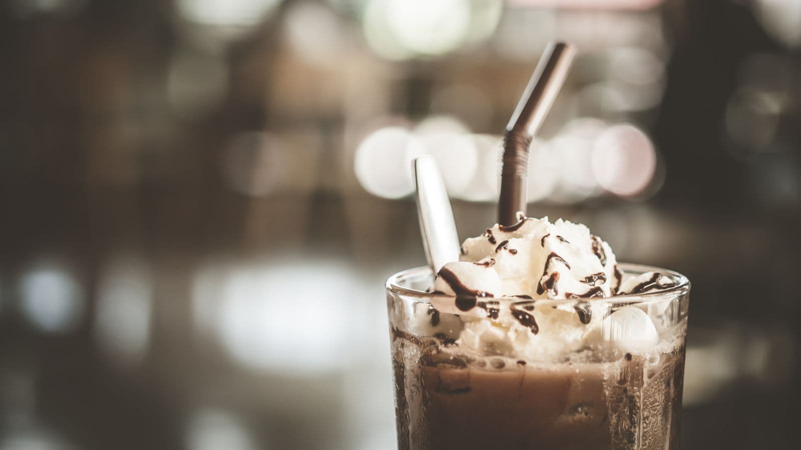 A glass of iced coffee topped with whipped cream and chocolate drizzle, featuring two straws. The background is softly blurred with circular bokeh lights.