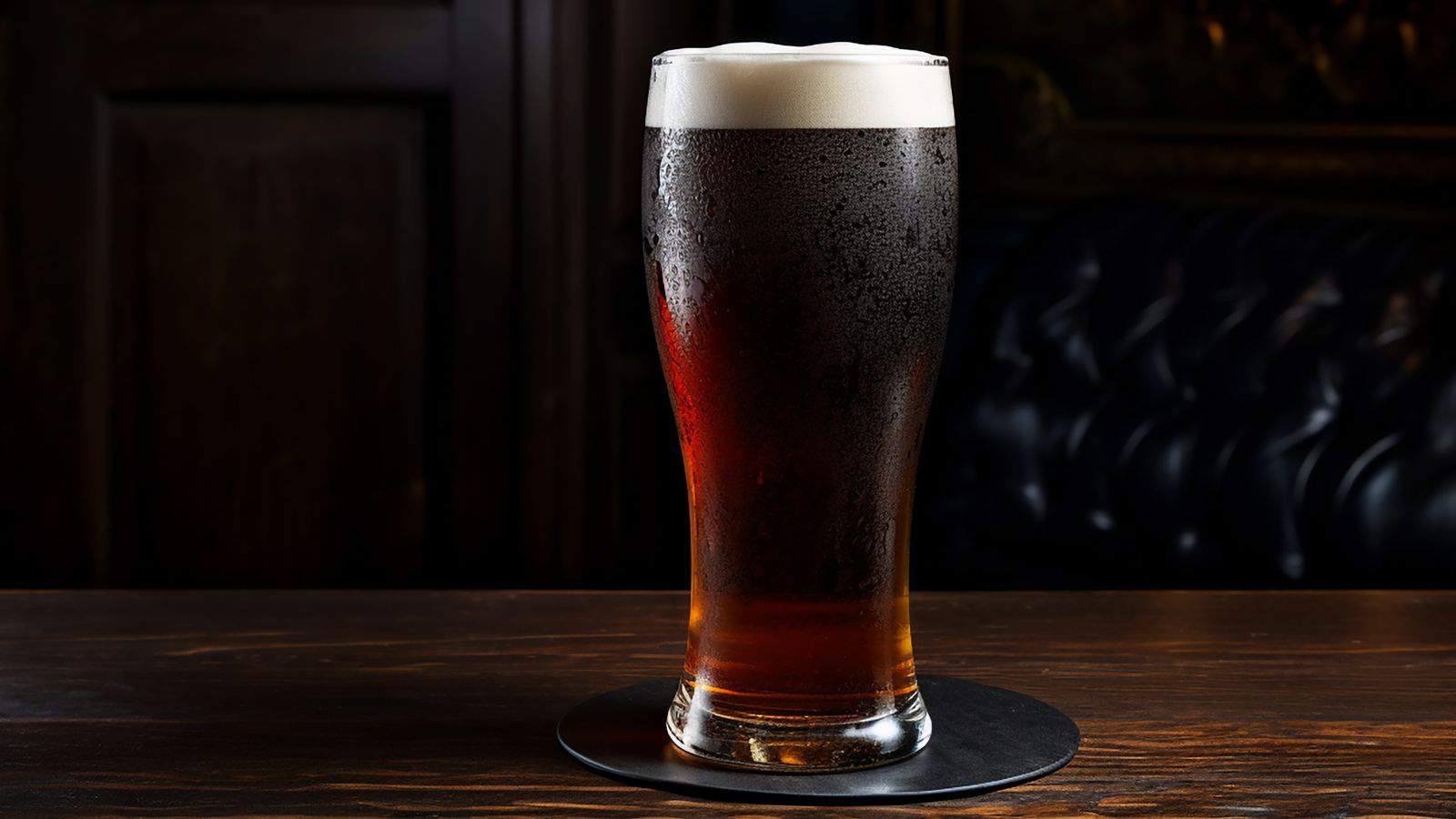A tall pint glass filled with dark beer sits on a black coaster on a wooden table. The beer has a light frothy head, and condensation is visible on the glass. The background is dimly lit, suggesting a cozy pub or bar setting.